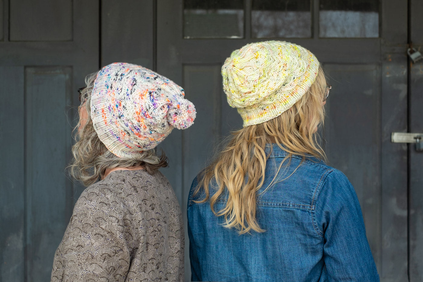 Jaime and Amber standing back to back, both wear handknits hats that are white with neon speckles, amber's is yellow speckles and Jaime's is coral, purple and blue speckles. The hats have a textured stitch.