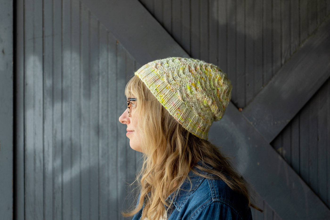 Side view closeup of Amber in her handknit hat, textured stitches in white yarn speckled with neon yellow, gold, and coral flecks here and there. 