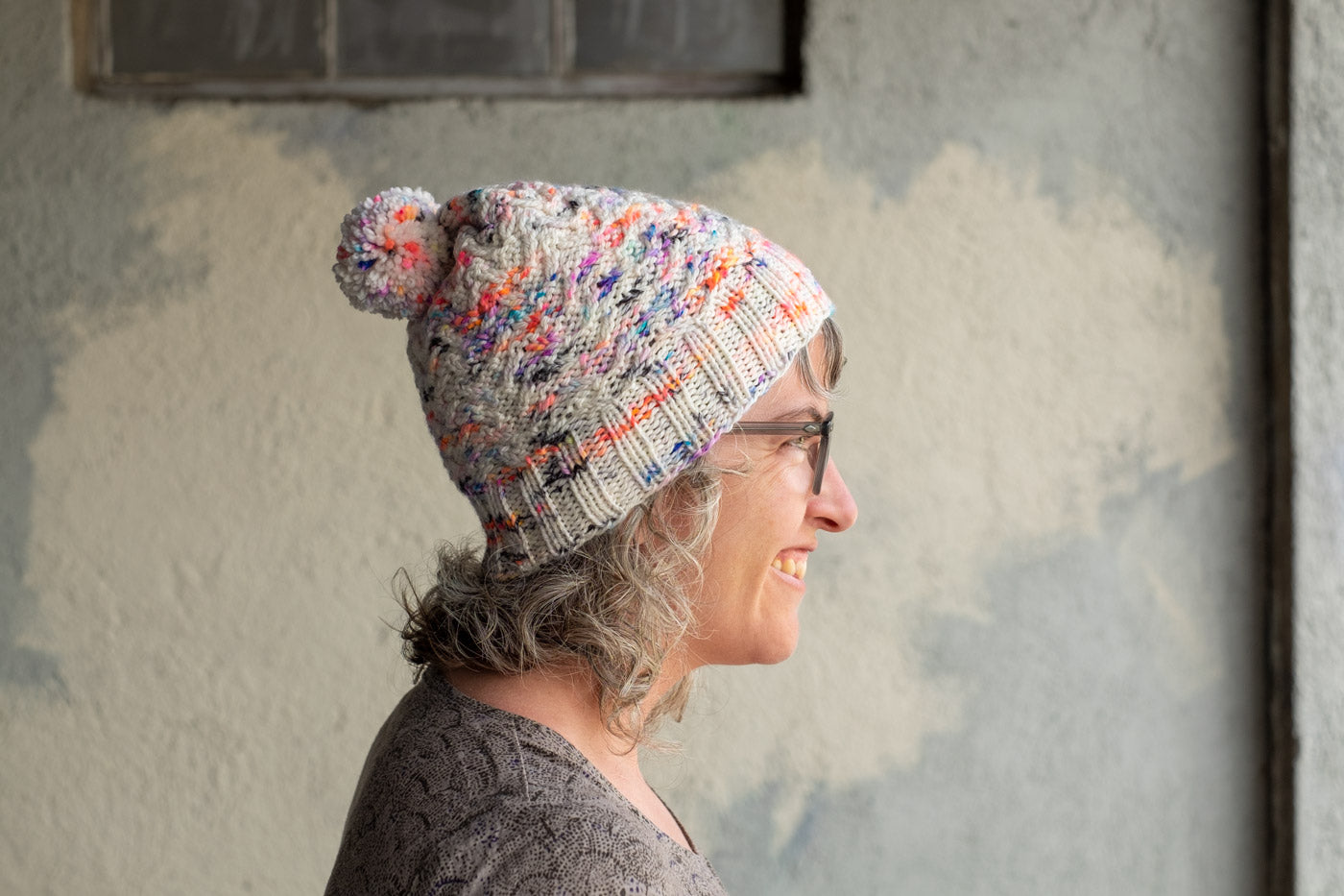 Side view closeup of Jaime in her handknit hat, textured stitches in white yarn speckled with coral, purple and blue. There is a pom-pom on top made from the same yarn.