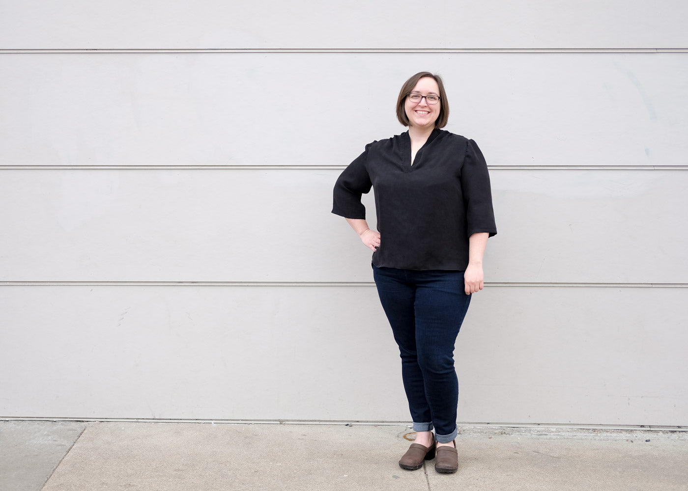 Woman wearing a drapey black blouse in front of a grey wall. 