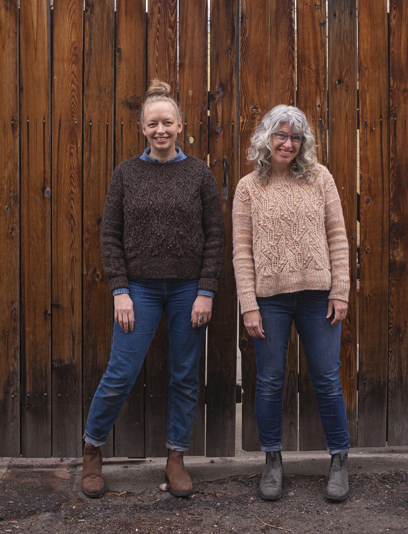 Amber and Jaime stand outside in front of a tall wooden fence on a rare damp day in Denver. They both wear their finished Atlantica pullovers: Amber's in a deep brown marl, and Jaime's in a pale peachy marl.