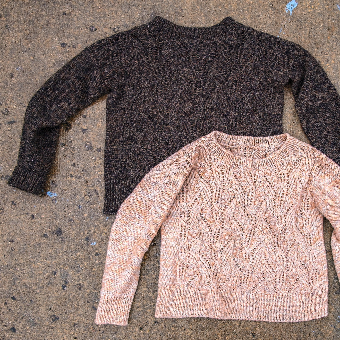 Two intricately textured sweaters lay on pavement, damp with spring rain. One is a dark brown marl, and one is pale peach marl. They both feature lace, bobbles, and cables knit in a laceweight Icelandic yarn. 