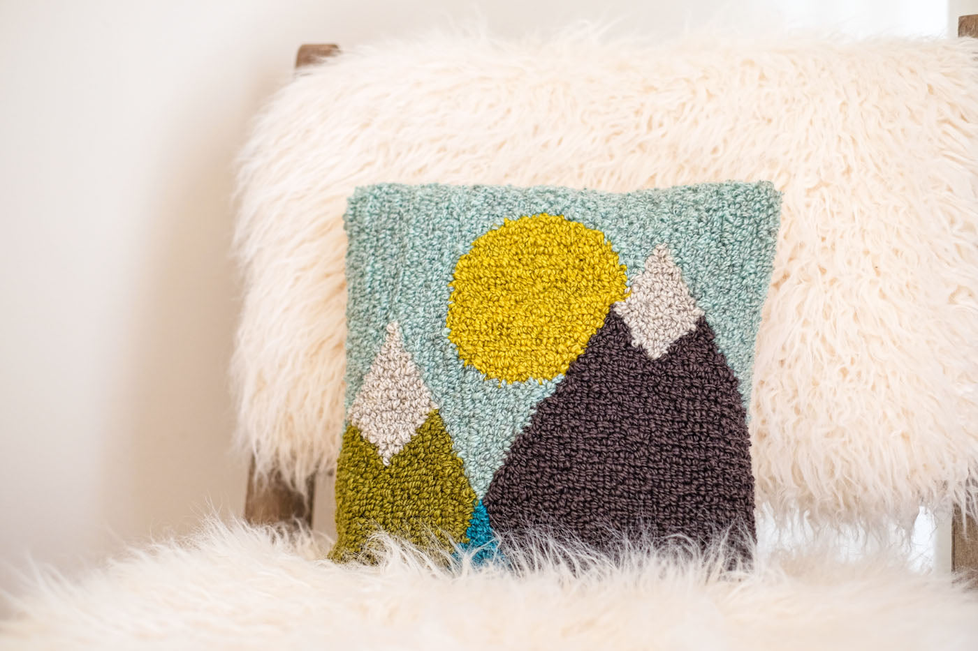 A punch needle pillow with geometric mountain and sun design. It is made in hues of green, blues, yellow, and grey, and is sitting on a plush sheepskin chair.