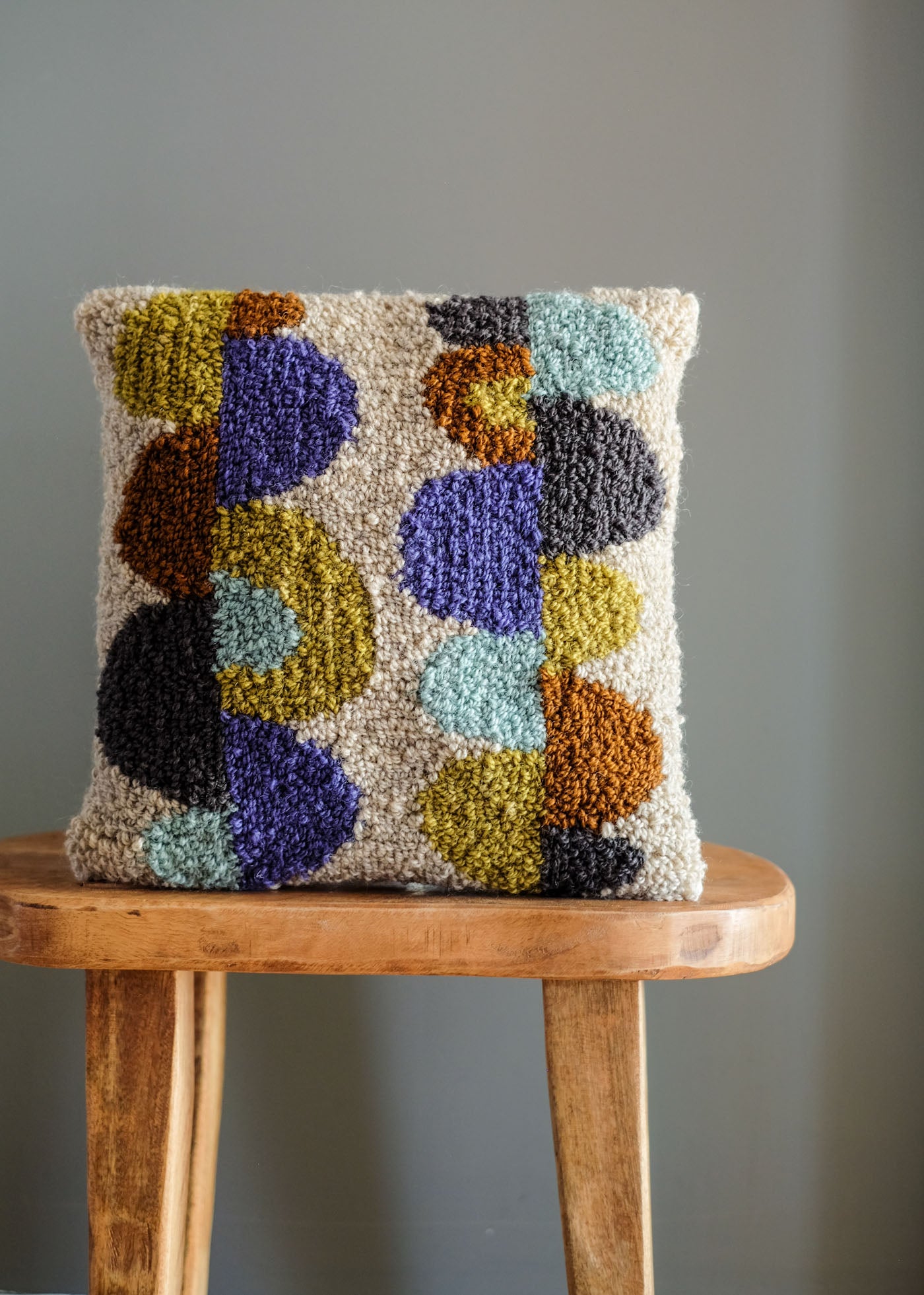 An abstract, modern punch needle pillow in hues of copper, grey, olive, pale turquoise and lavender. 