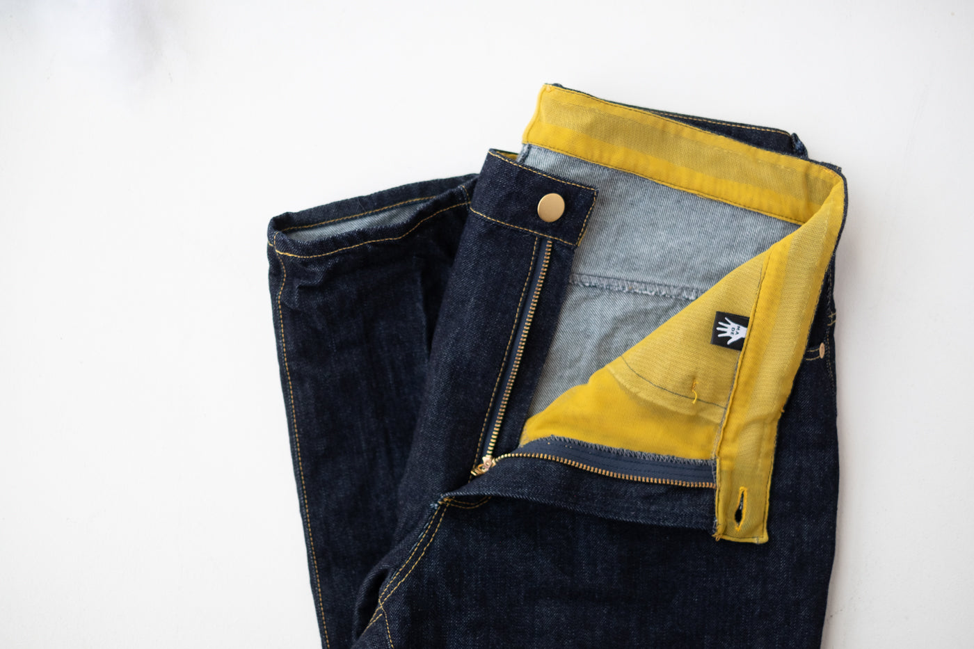 Demin jeans laying flat, folded on a white background.  The zipper is unzipped, folded back to show the yellow mesh lining with a custom label.