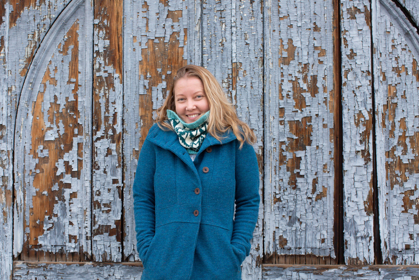 Danielle agains a peeling grey wooden backdrop.  Danielle is in a dark sky blue peacoat with her hand in her pockets.  With a white, turquoise and teal cowl around her neck.