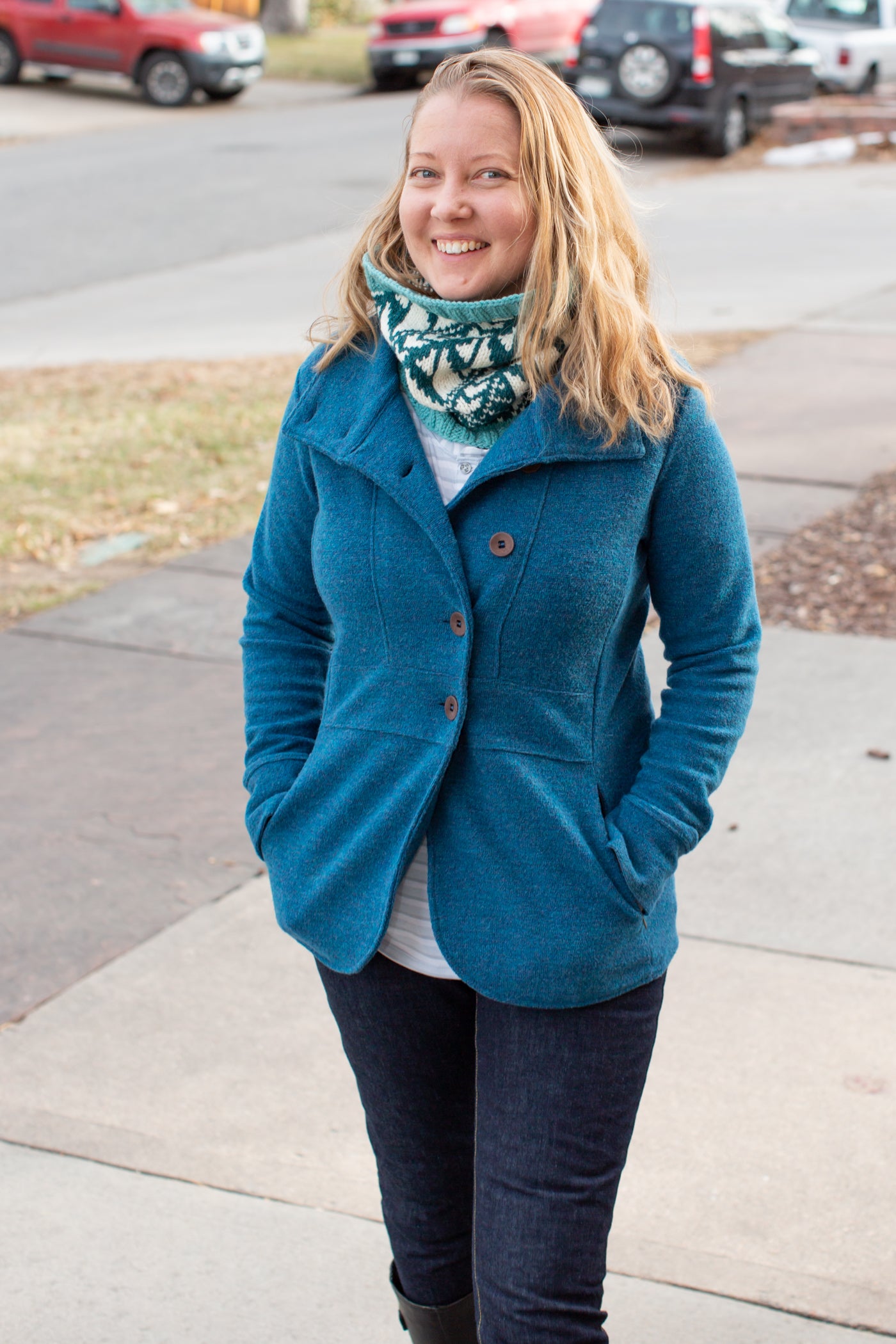 Danielle walking down a city sidewalk smiling.  Danielle is in a dark sky blue peacoat with her hand in her pockets.  With a white, turquoise and teal cowl around her neck.