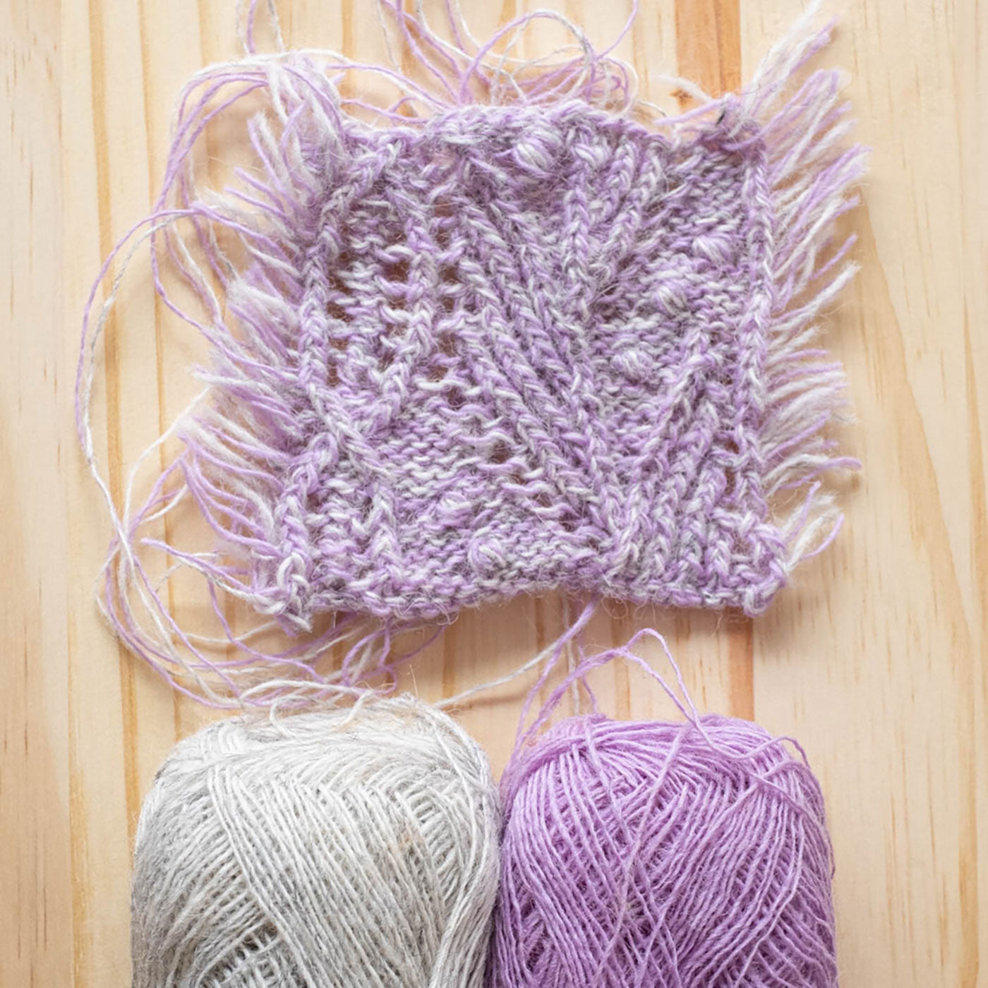 A knitted swatch for the Atlantica Pullover in a marl of two colors held together: Lavender and Light Ash Heather— lays on a background of light unfinished pine wood. The swatch features a delicate cabled lace. Two yarn balls in Lavender and Grey peek into the bottom of the picture. 