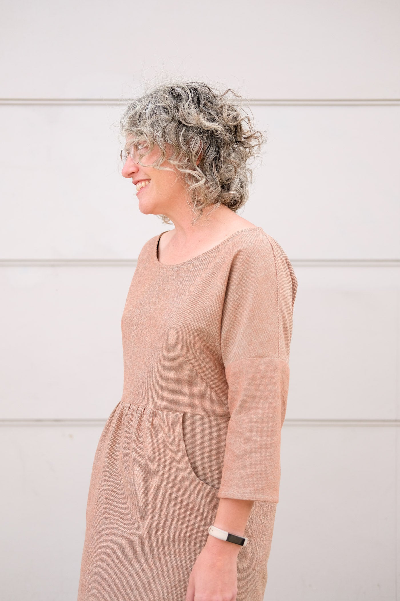 Close up of woman in a pale brown, subtly textured dress. She stands outside in front of an off white wall, and is turned to the side.