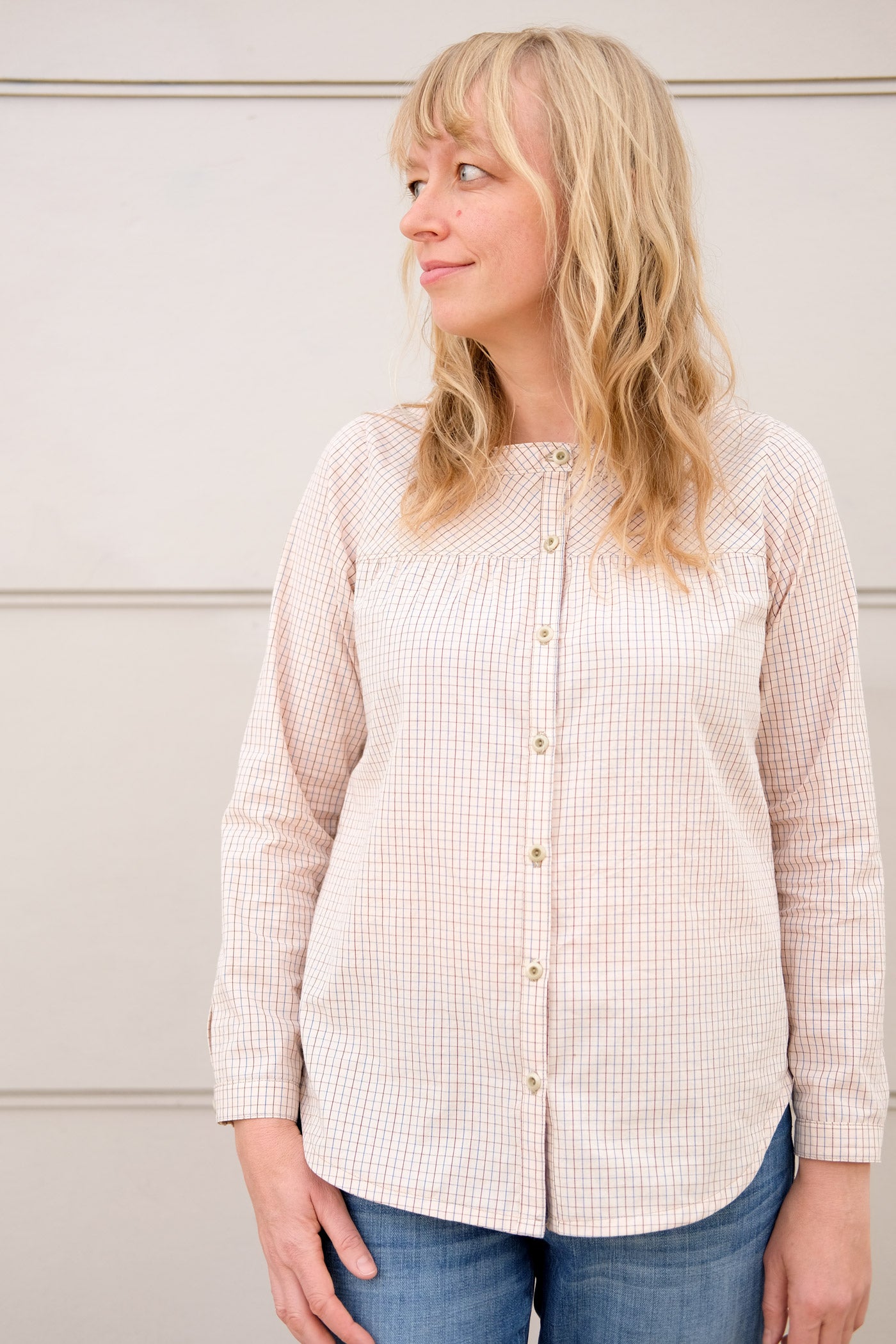 Woman wearing a button up shirt in a very pale peach, subtle plaid, standing in front of an off-white wall. The shirt is a gently gathered at the yoke, long sleeved and has a minimal band collar.  