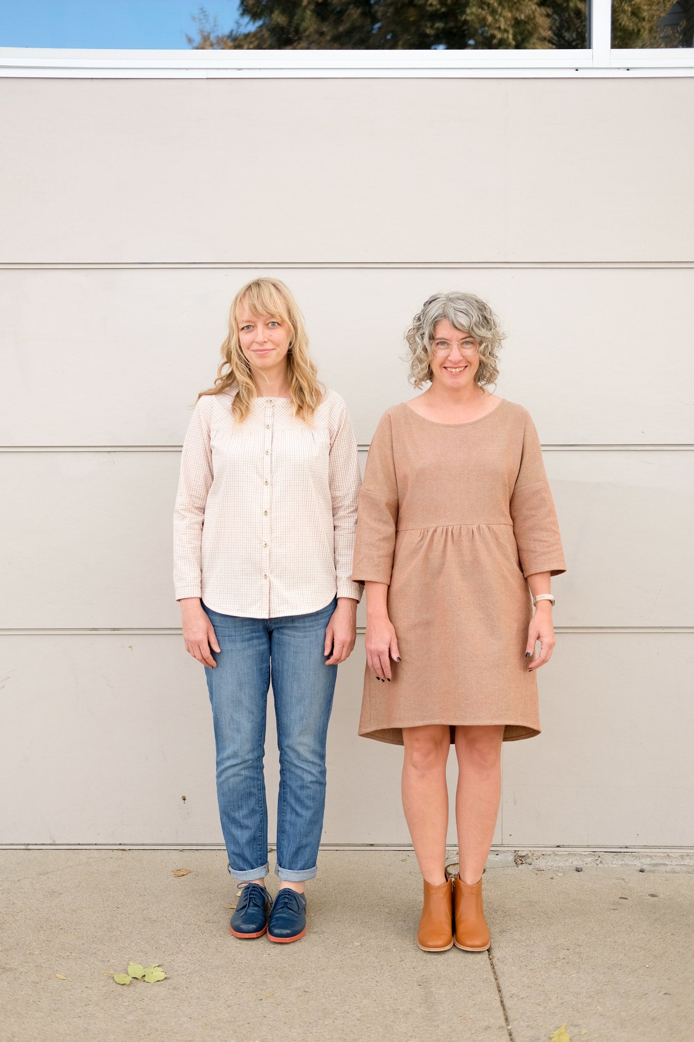 Two women standing in front of an off-white wall outside. One wears jeans and a light plaid button up shirt, and the other wears a light brownish knee-length dress with 3/4 sleeves and a gathered waist with pockets.