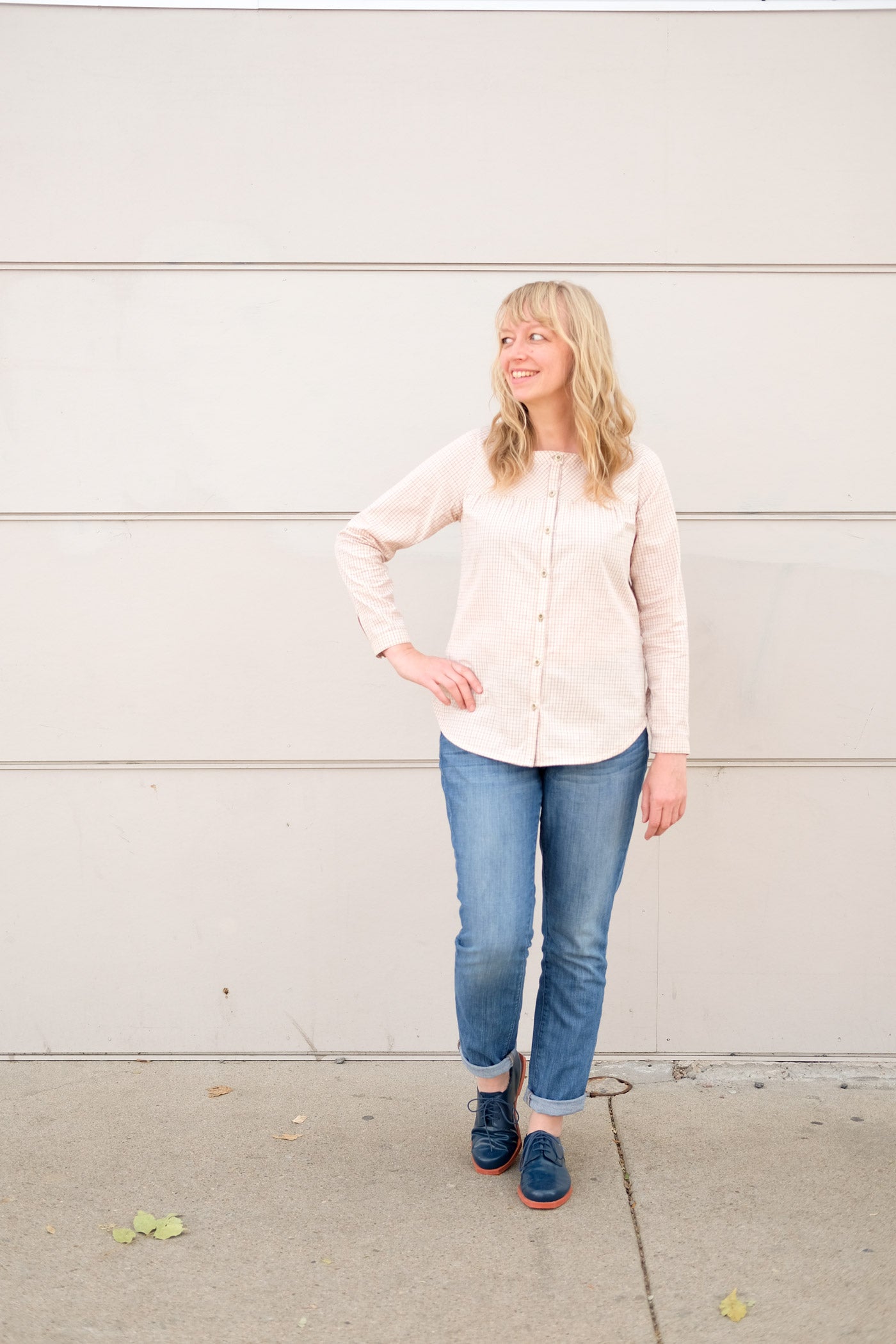Woman standing in front of an off white wall in jeans and a very pale peach button up shirt. She smiles and looks off to the left with one hand on her hip.