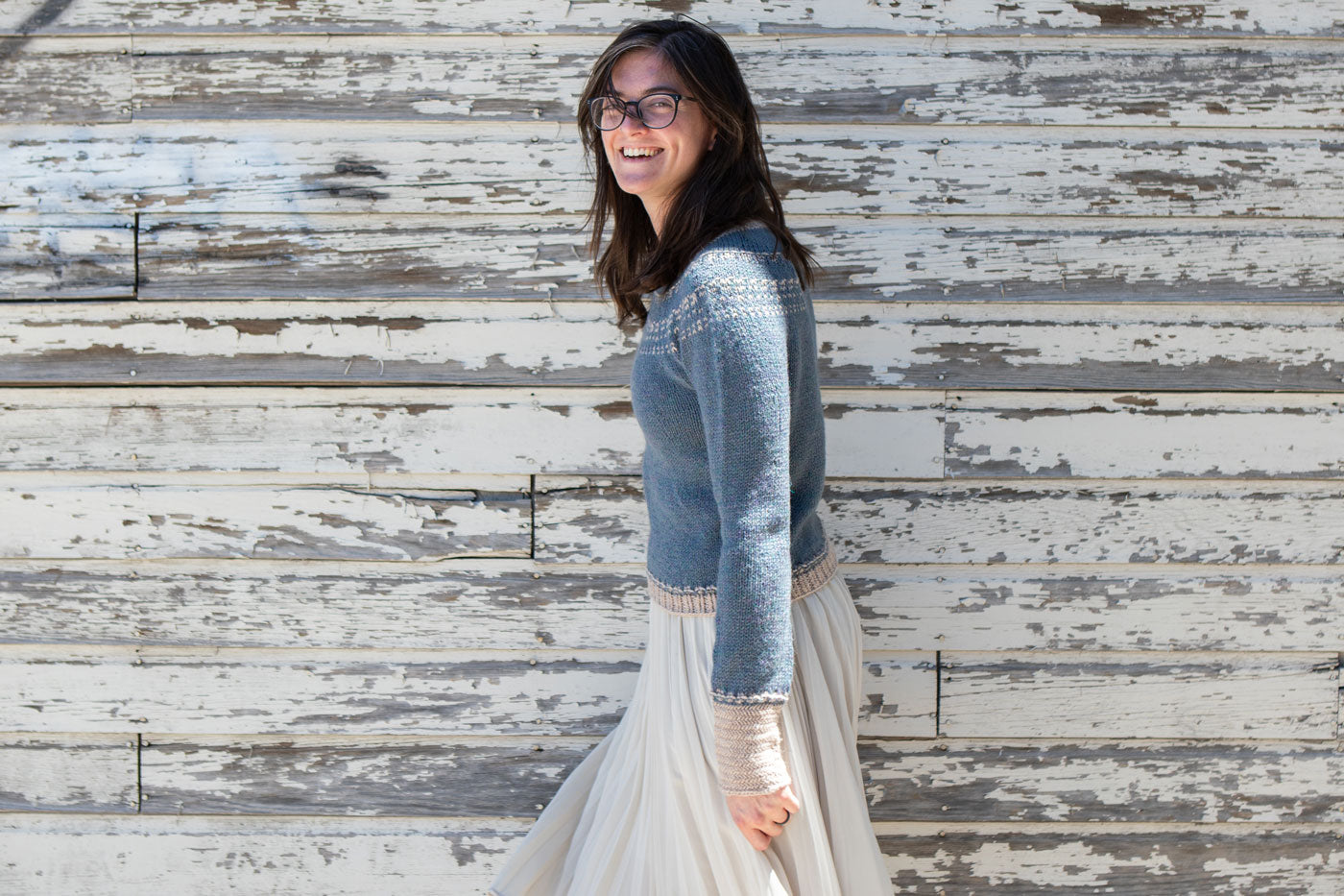 Flavia stands in front of a white washed wall while wearing her Sprout Pullover paired with her white skirt. Her side profile faces the camera while smiling at the camera