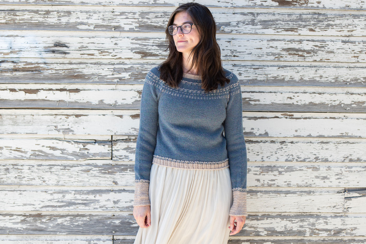 Flavia wears her Sprout Pullover, paired with a white skirt, she stands in front of a white fence while looking away.