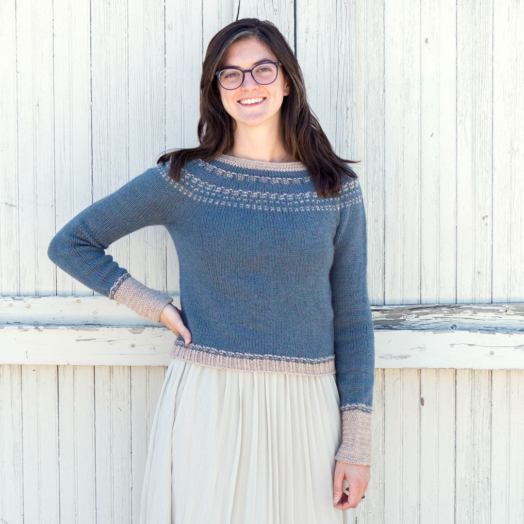 Flavia stands in front of a tall white fence. She is wearing her Sprout Pullover and a long white skirt. She has her right hand on her hip while she is smiling.