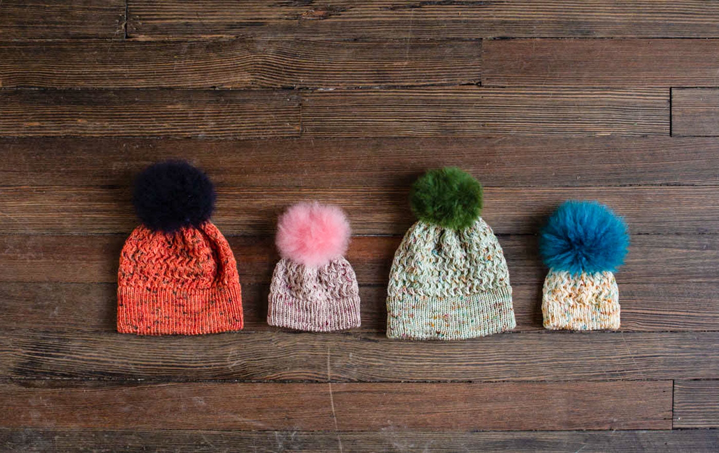 Four hand knit hats lay in a row on a dark wood background. They are made in a variety of sizes from newborn to adult, and a variety of hues of speckled, hand-dyed yarn. Each one is topped with a soft, alpaca fur pompom in a coordinating or contrasting color: a large hat in fiery orange with deep navy speckles is topped with a dark navy pompom; a small pink and grey hat is topped with a pale pink pompom; a sage green hat is topped with an olive green pompom; a tiny newborn hat in speckles of beige and turquoise is topped with a vibrant turquoise pompom. 