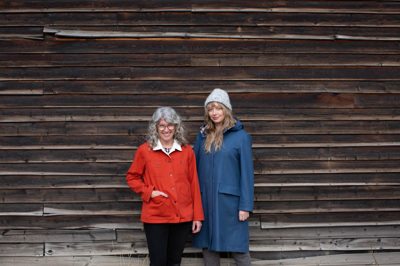 Amber and Jaime stand together outside against a weathered barnwood wall. Jaime wears her vibrant red Thayer Jacket with cream fleece lining showing at the collar. Amber wears her blue wool Hoodie Parka with a hand knit grey hat.