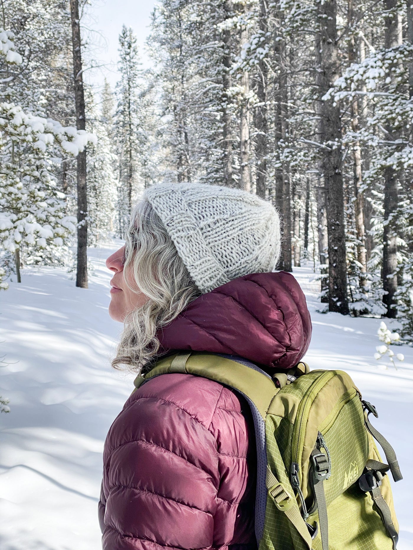 Jaime wears a pale grey, tweedy ribbed hat. She stands in a snowy forest of tall evergreens, keeping warm in a burgundy puffy jacket and olive hiking pack.