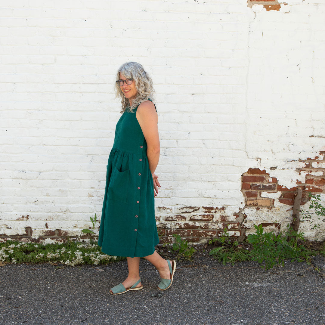 Jaime stands in front of a white brick wall wearing her Austin Dress in an emerald color. She stands sideways so that the button details on the side of the dress are visible. 