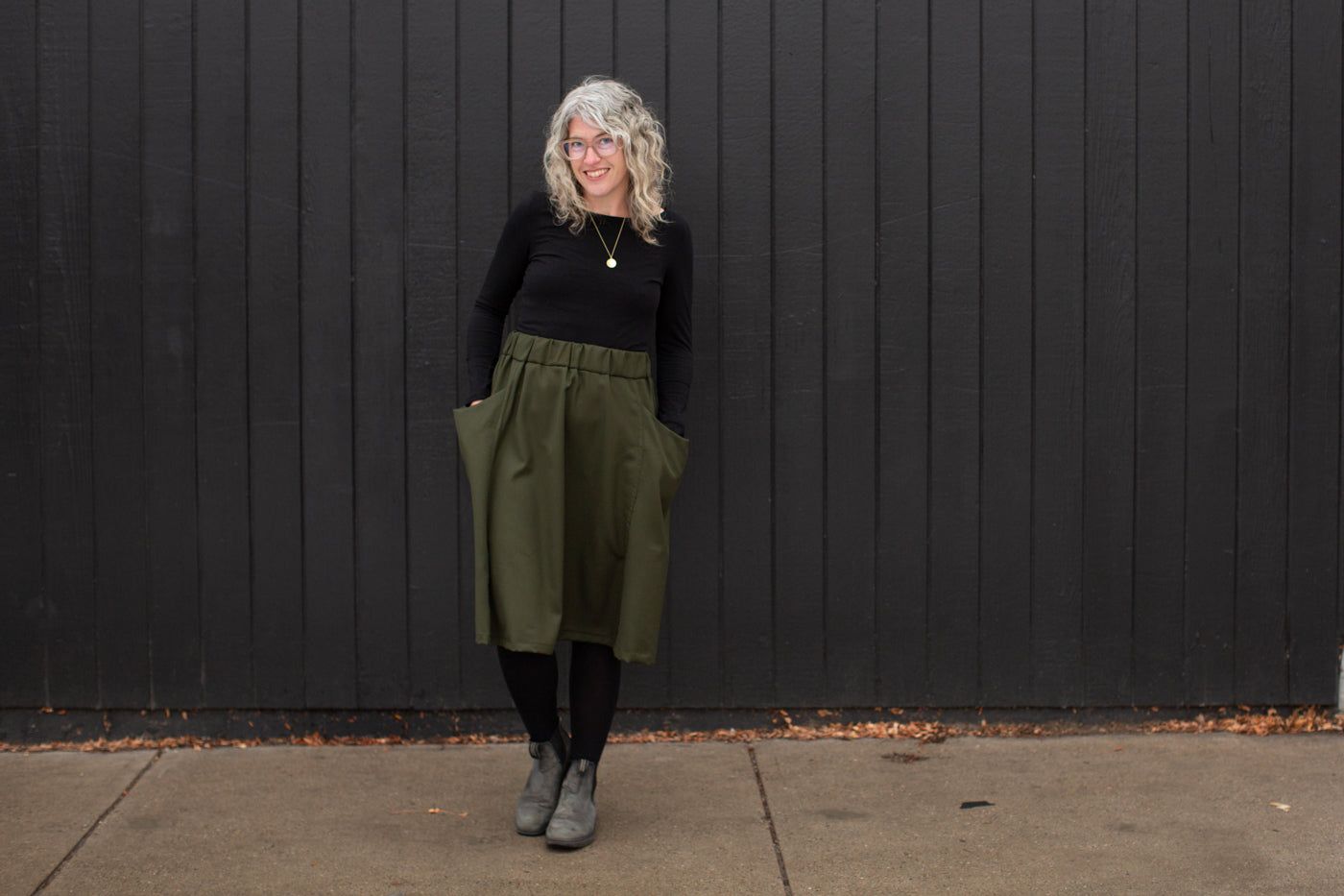 Jaime standing against a black wall looking at the camera smiling.  Jaime is wearing a black long sleeve shirt, olive green skirt, black leggings and black shoes.