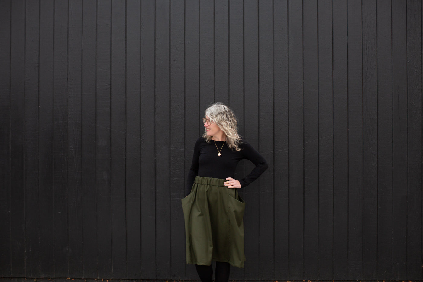 Jaime standing against a black wall in a black long sleeve and olive colored skirt with her hands in her pockets looking off to the right.