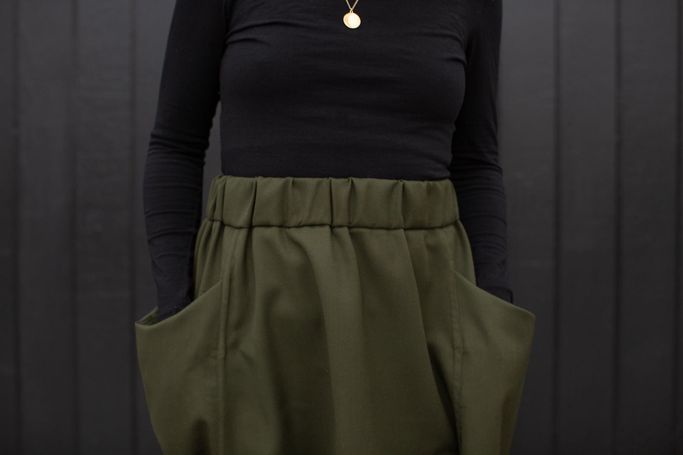 Close up of Jaime standing against a black wall, wearing a black long sleeve shirt with her hand in the pockets of an olive green skirt.