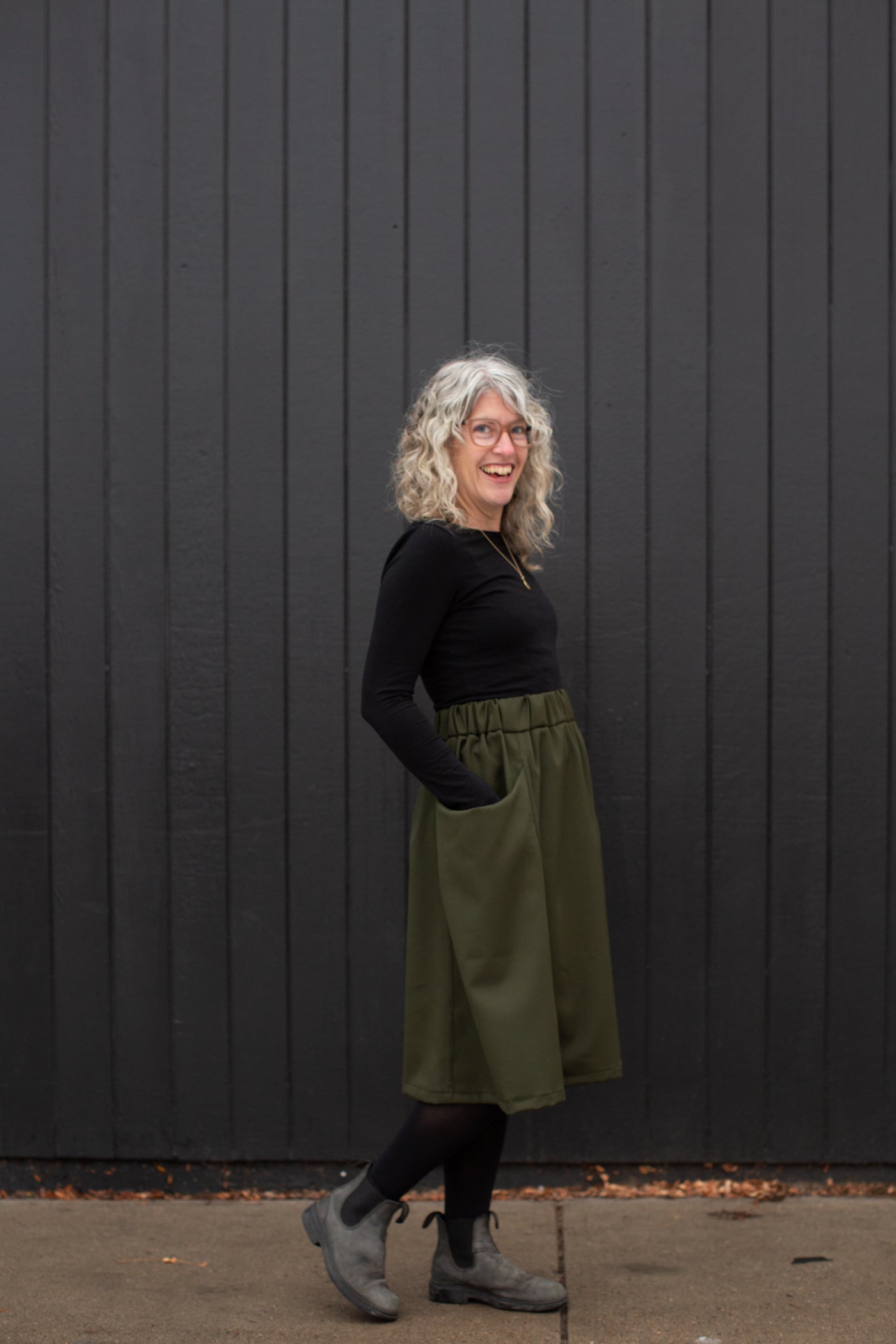 Jaime standing against a black wall in a black long sleeve and olive colored skirt with her hands in her pockets looking at the camera smiling.