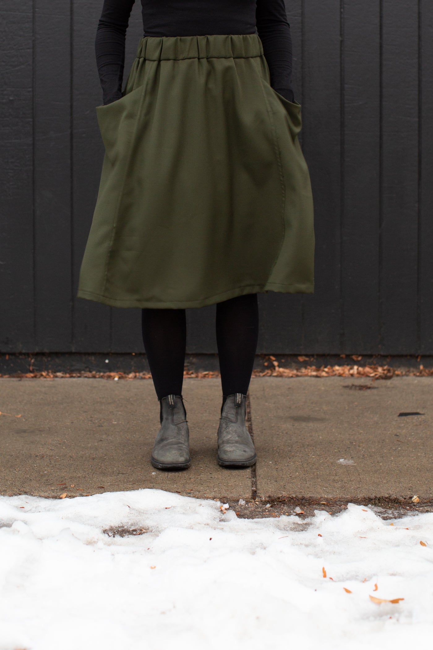 Mid waist to feet image of Jaime standing against a black wall, wearing an olive green skirt, black leggings and shoes with her hands in the pockets.  Jaime is standing on a sidewalk with snow in the foreground.