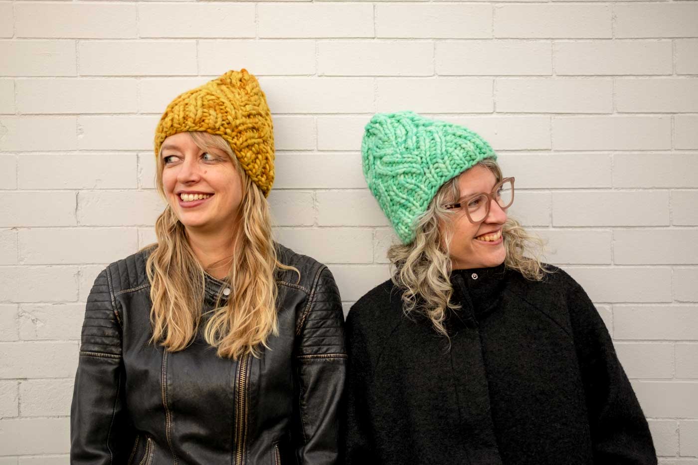 Amber in a yellow Neighborhood Holiday hat and Jaime in a mint green one, standing against a white brick wall.
