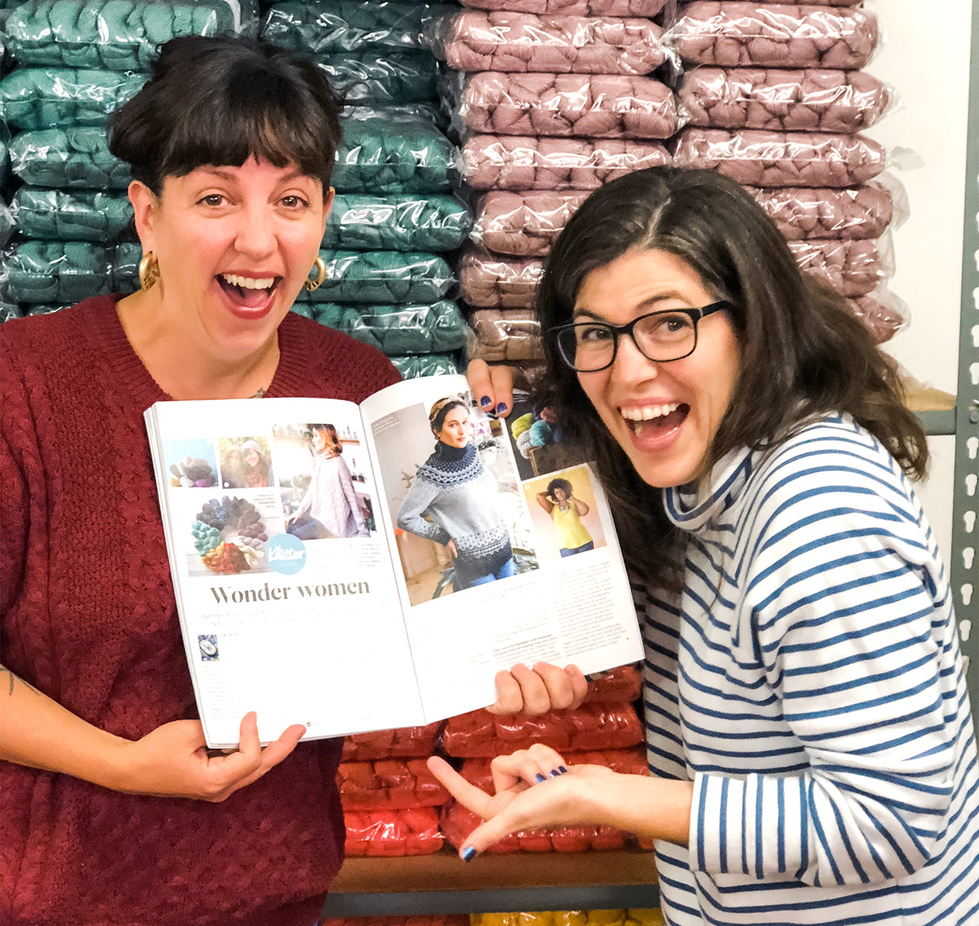 This is an image of two women holding a book open laughing in front of a wall of yarn. 