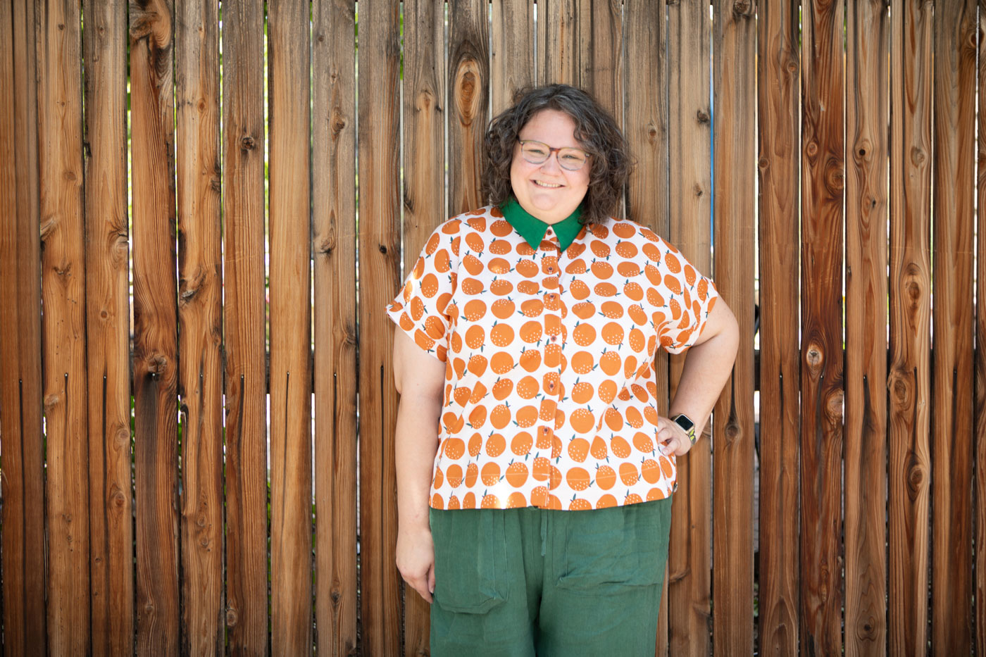 Kim wears her orange printed Kalle Shirt. She stands in front of a brown fence with with her right hand on her hip. She waits a pair of emerald green pants that matches the green collar of the shirt.