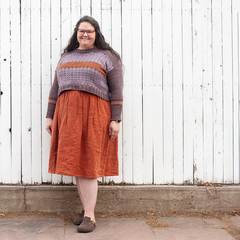 This is an image of Kim wearing her Mozaika Sweater in Corrie Worsted Quartz Fume, Rust, and Abalone while standing in front of a tall white fence. 