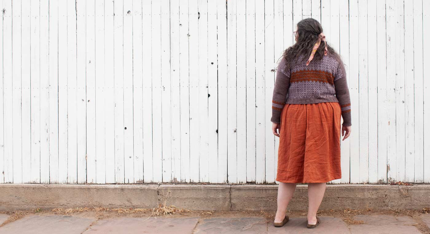 This is an image of Kim with her back to the camera wearing her Mozaika sweater in Corrie Worsted Quartz Fume, Rust, and Abalone in front of a tall white fence. 