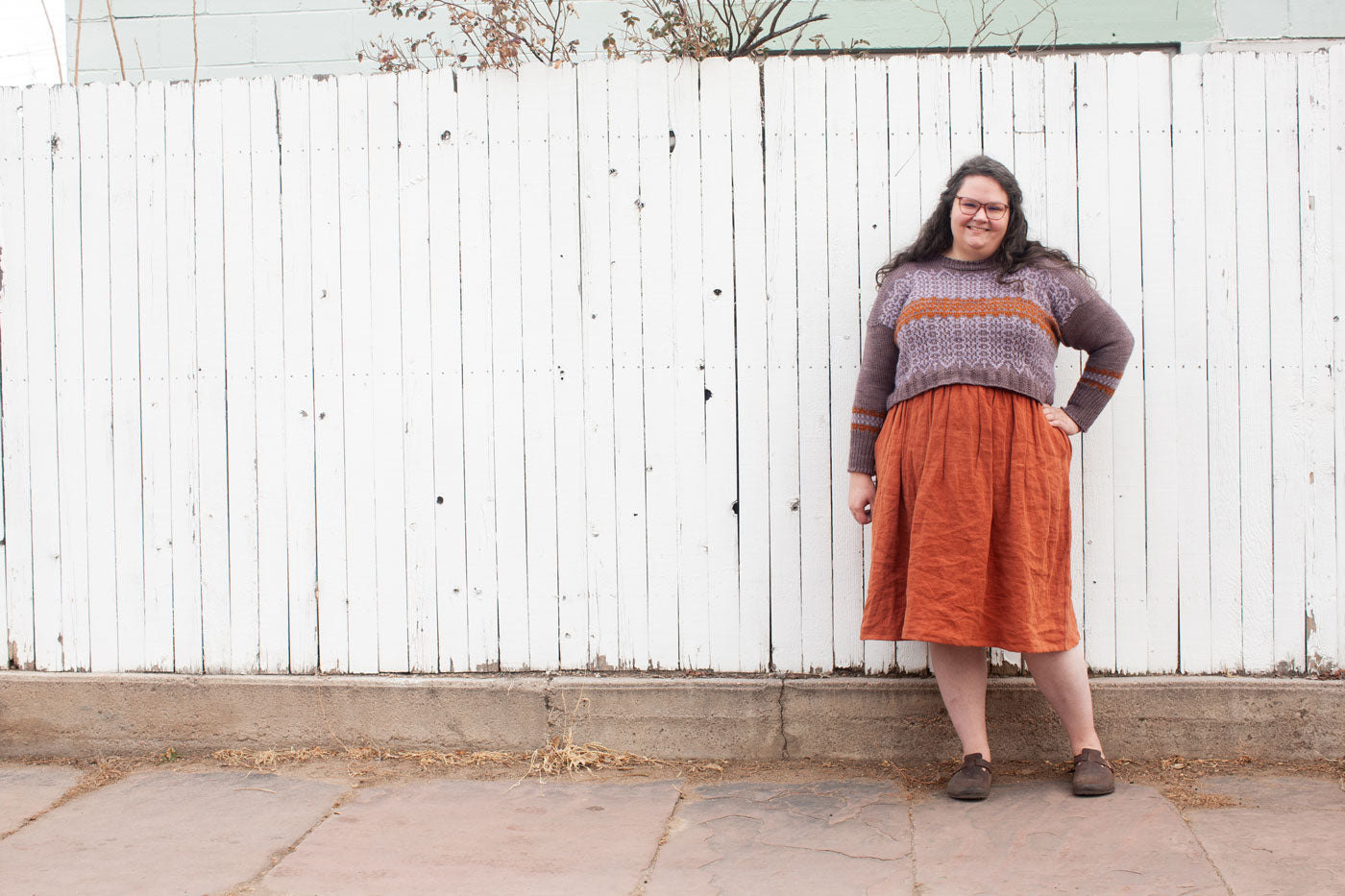 Kim stands in front a tall white fence. She is wearing a pink sweater and a rust midi skirt. 
