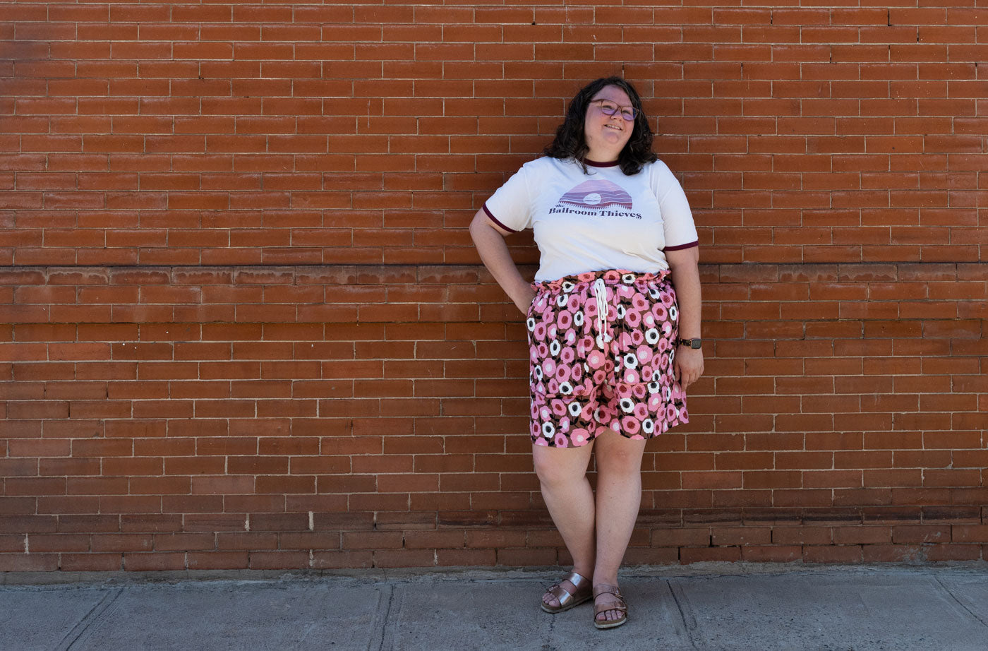 Kim wears her Soline Shorts made from a bright pink floral barkcloth fabric. She is wearing a white graphic t-shirt and stands in front of a brick wall.  