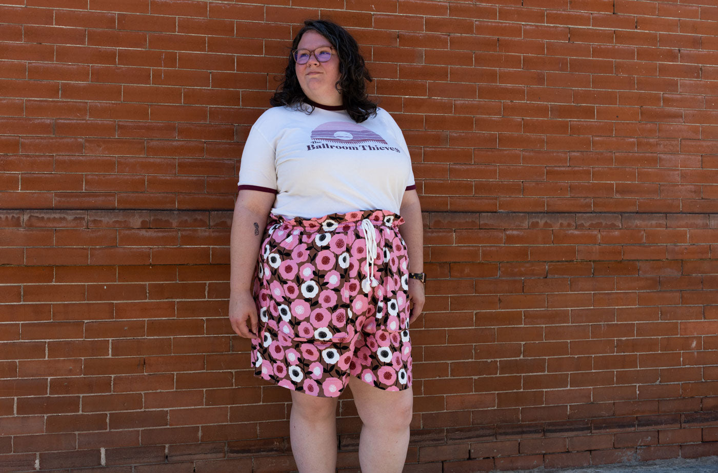 Kim wears her Soline Shorts made from a bright pink floral barkcloth fabric. She is wearing a white graphic t-shirt and stands in front of a brick wall.  