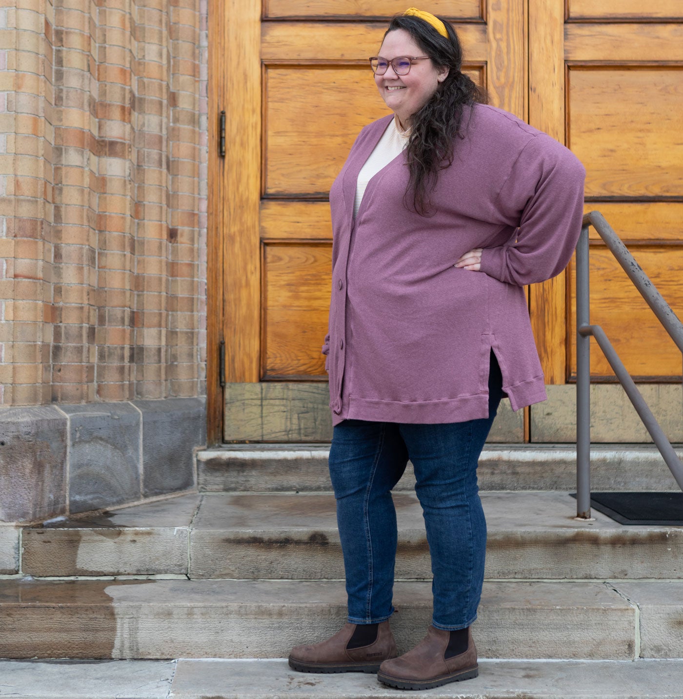 This is an image of Kim wearing her Marlo Sweater in rayon cotton modal sweater knit in Rose brown. She is standing on steps in front of a light brown door. She is also wearing a yellow headband, a white shirt underneath her cardigan, and blue jeans. 