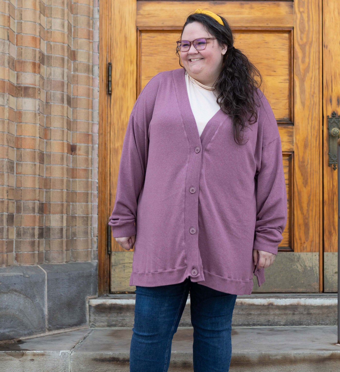 This is an image of Kim wearing her Marlo Sweater in rayon cotton modal sweater knit in Rose brown. She is standing on steps in front of a light brown door. She is also wearing a yellow headband, a white shirt underneath her cardigan, and blue jeans. 