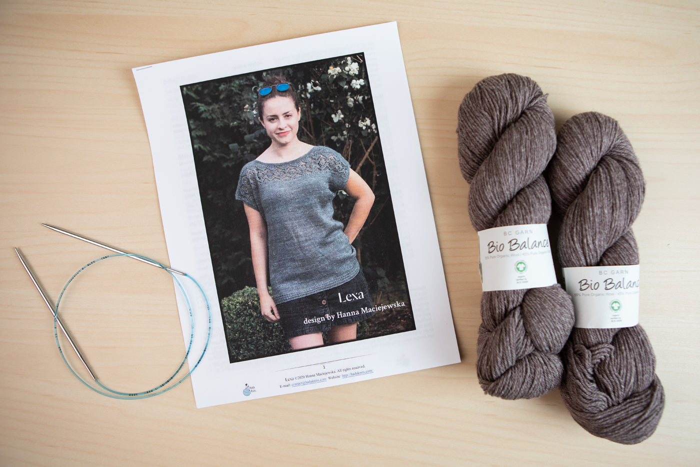 Kim’s chosen Lexa knitting pattern, Her charcoal-hued BioBalance yarn and a set of knitting needles lay on a light wood background.