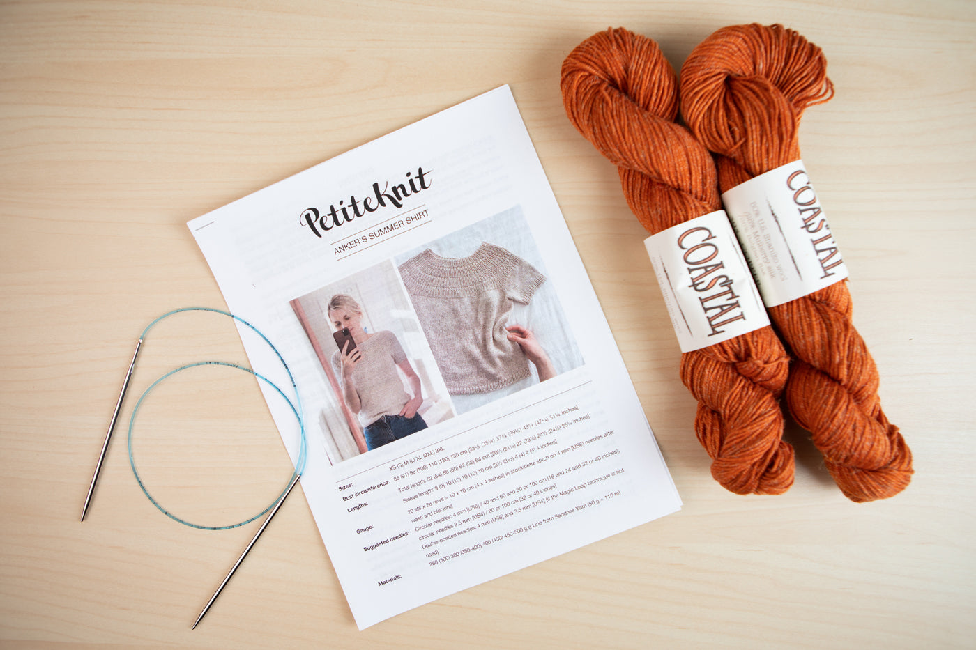 Jaime’s chosen PetiteKnit pattern, Copper-hued Coastal yarn and a set of knitting needles lay on a light wood background.