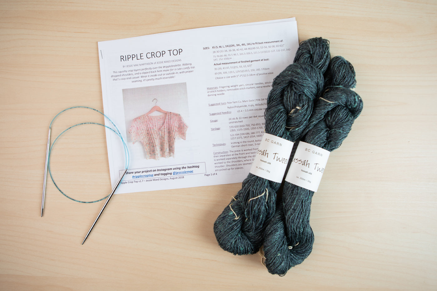 Marta’s chosen Jessie Mae pattern, and Tussah Tweed yarn in a deep muted teal, and a set of knitting needles lay on a light wood background.