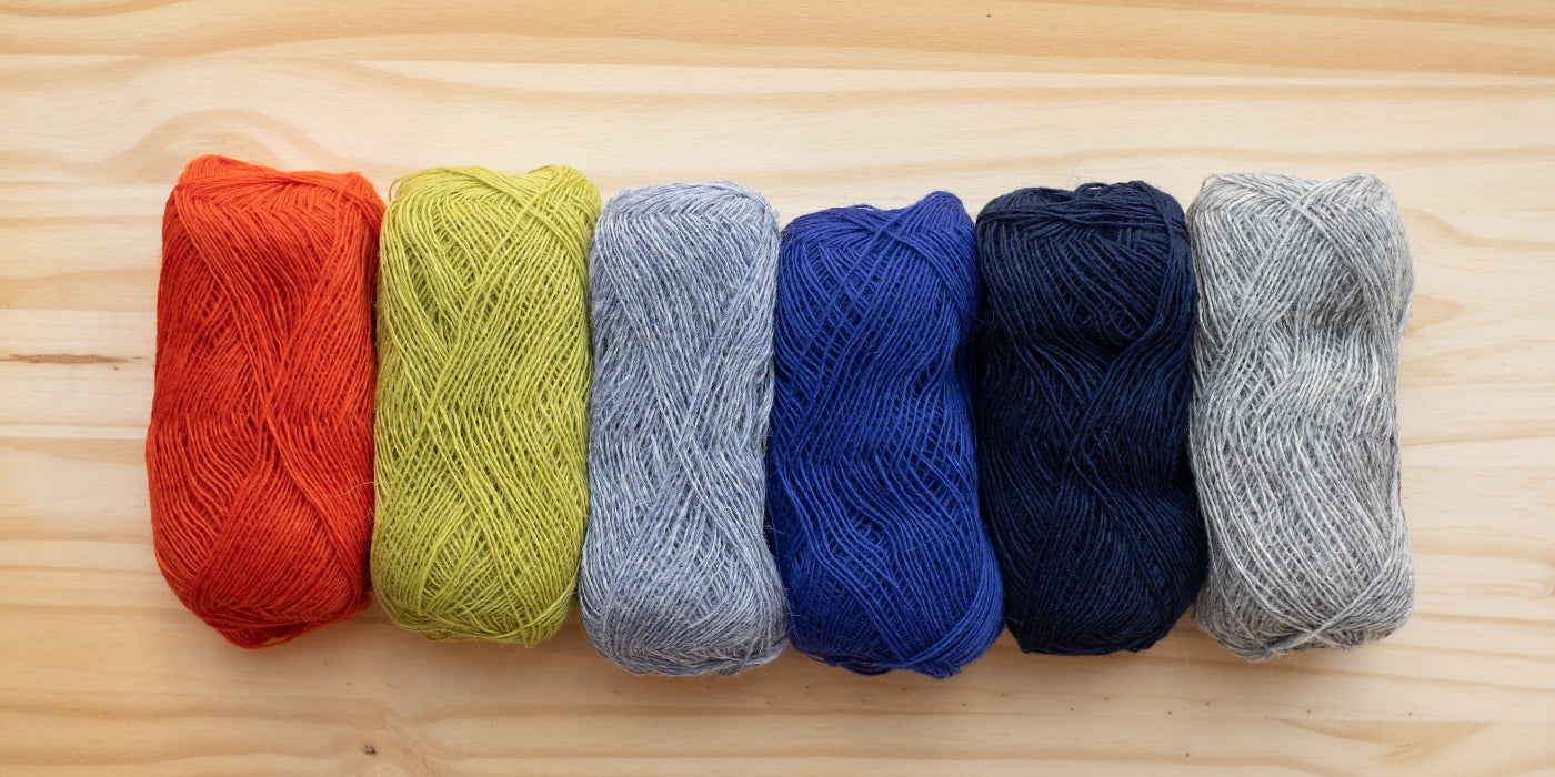 Lopi skeins in different colors lined up in a lay flat image with a birch wood background.  The colors from left to right are bright orange, light grass green, light blue, colbalt blue, navy and heather grey.