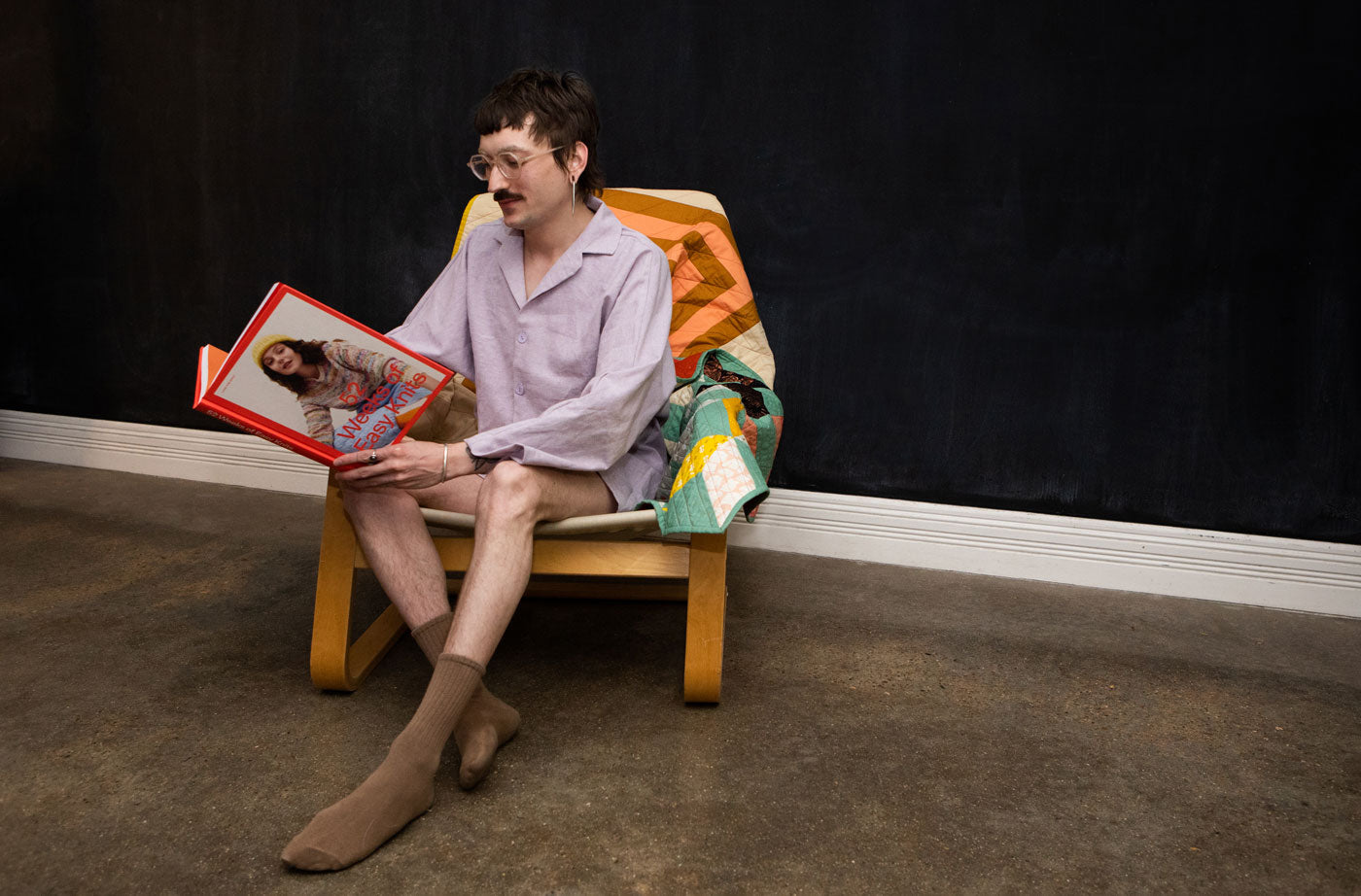 Leaf sits in an arm chair reading a book while wearing their Agnes PJs in Essex Linen lilac