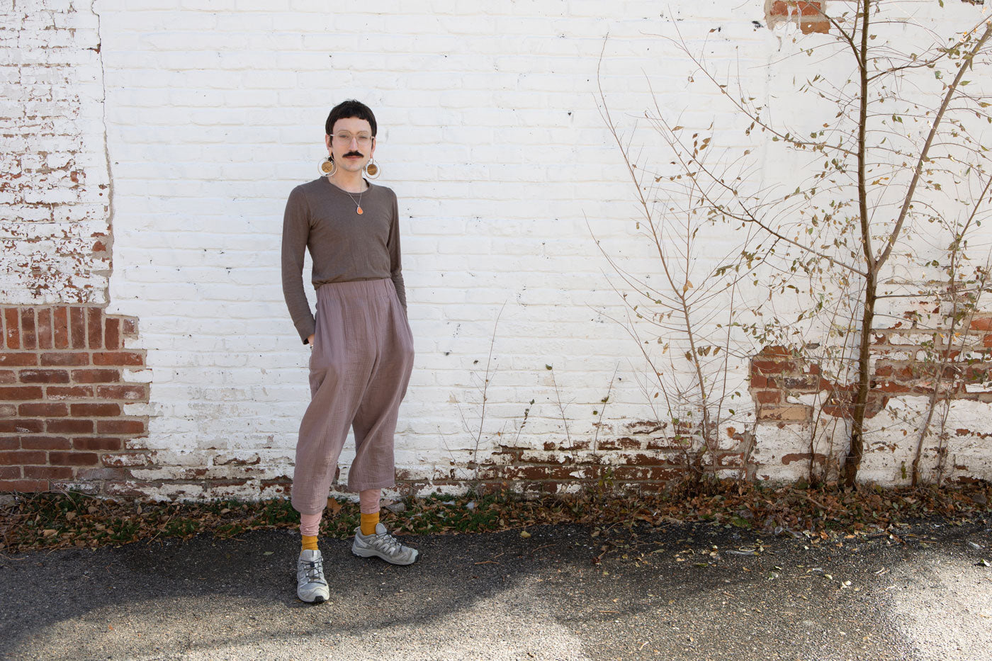 Leaf stands in front of a white brick wall. They are wearing a purple long sleeve shirt and flowly violet pants.