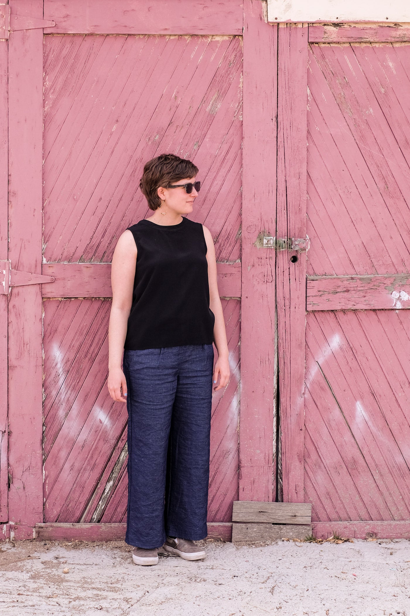 Woman standing outside a weathered red barn door, wearing sunglasses, a black tank top and wide legged indigo linen trousers.