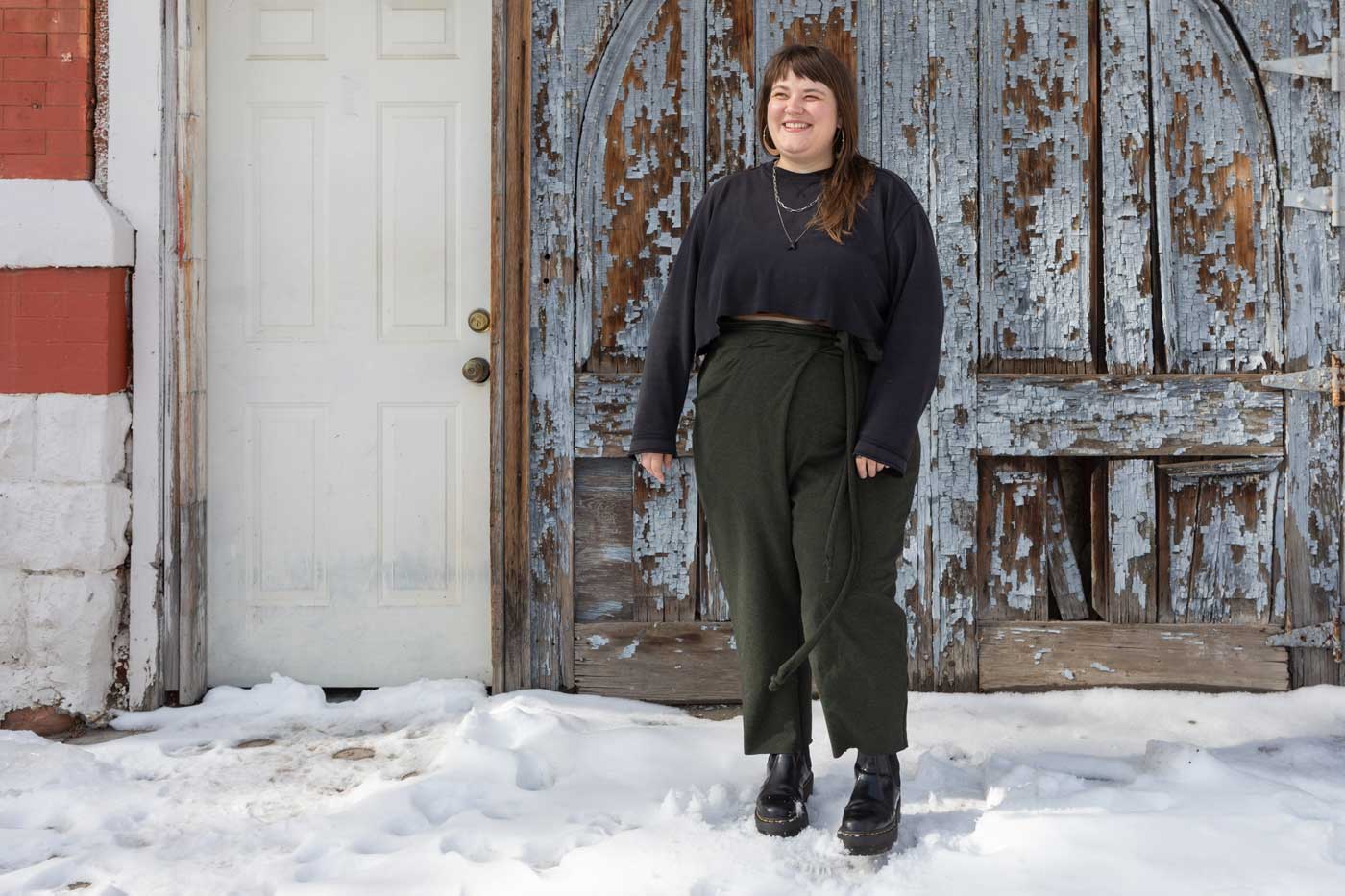 This is an image of Marta wearing her Papao Wrap Pants in Bamboo Cotton Stretch French Terry - Heather Forest, she is also wearing a dark grey crop sweatshirt while standing in the snow in front of a rustic wooden door.