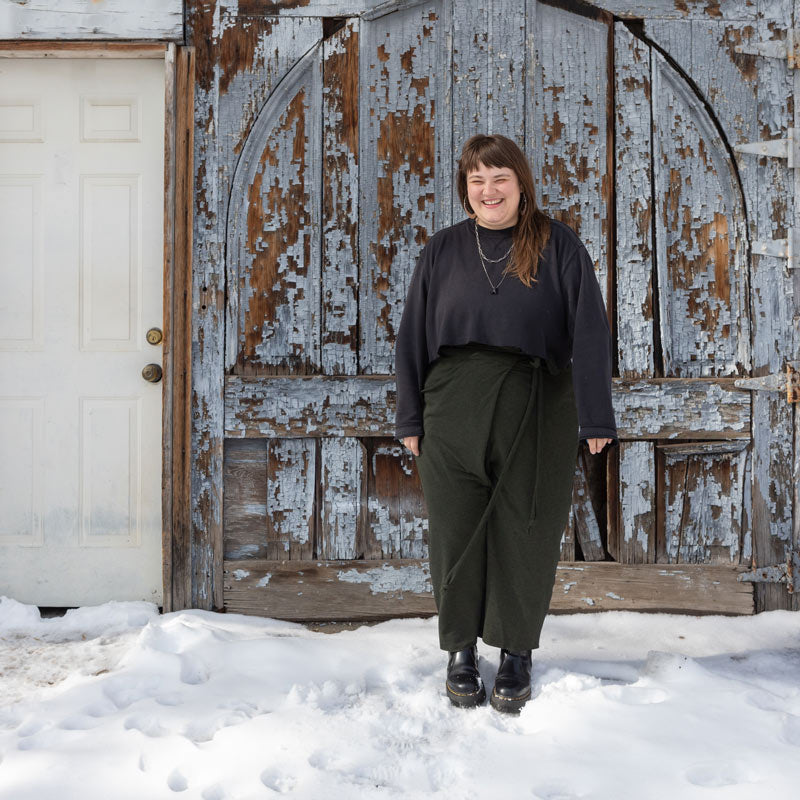 This is an image of Marta wearing her Papao Wrap Pants in Bamboo Cotton Stretch French Terry - Heather Forest, she is also wearing a dark grey crop sweatshirt while standing in the snow in front of a rustic wooden door.