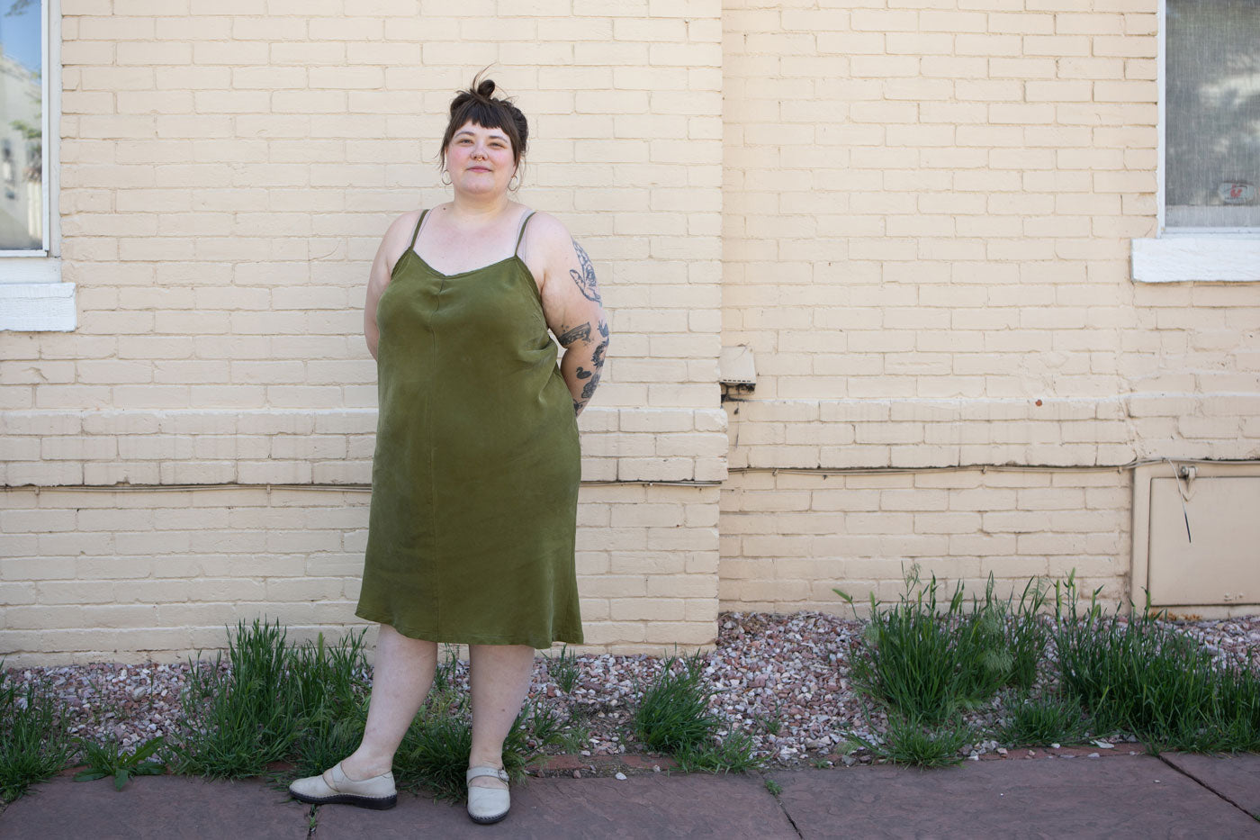 Marta stands in front of a light yellow brick wall wearing her Saltwater Slip dress in the color moss. Her hair is up in a a bun.