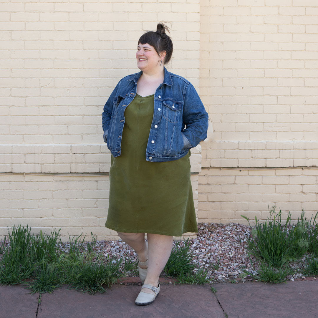 Marta is wearing her Saltwater Slip dress in the color moss. She is wearing a jean jacket and is standing in front of a light yellow brick wall. 