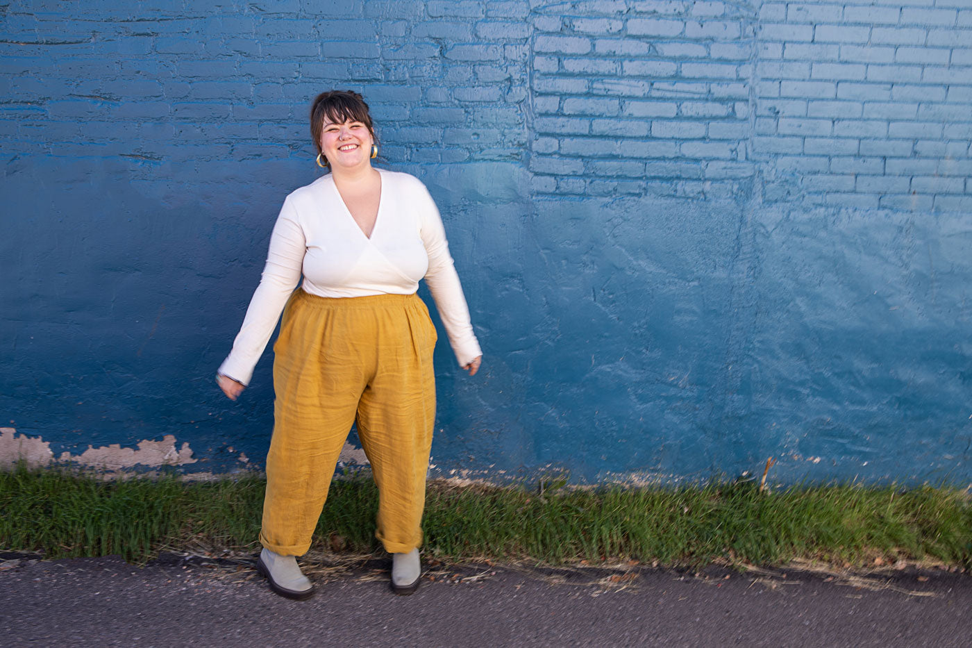 Marta is standing in front of a blue brick wall. She is wearing a long sleeve white wrap top and yellow top. 